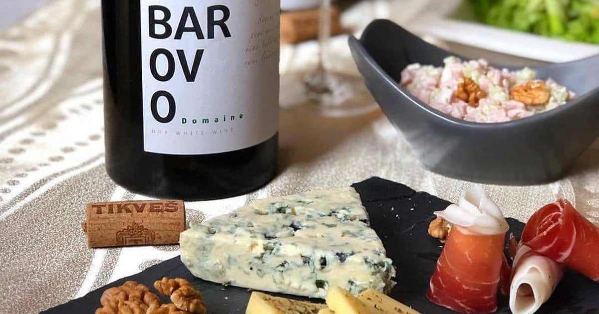 A bottle of Tikveš Barovo white beside a slate board of blue cheese, hard cheese, walnuts and cured ham.