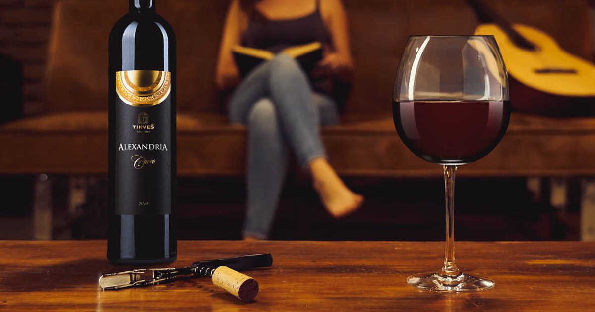 A bottle of Tikveš Alexandria Cuvée red on a wooden table with a pulled cork, a corkscrew and a full glass.