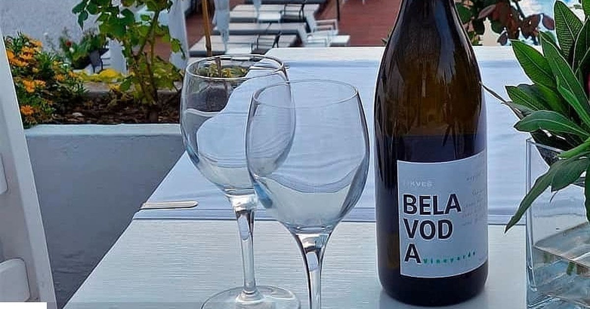 A bottle of Tikveš Bela Voda white with two empty glasses on a terrace table above the sea.