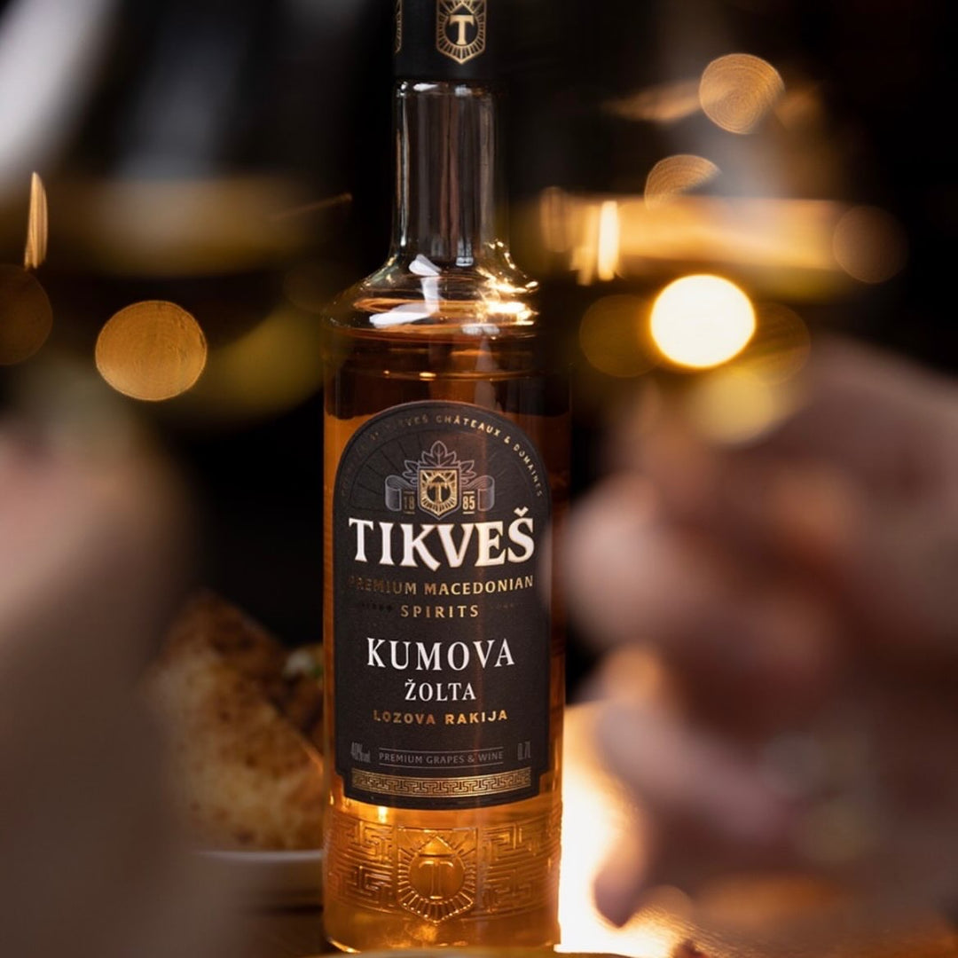 A bottle of Kumova Žolta grape rakija on a table in warm evening light, with glasses raised either side