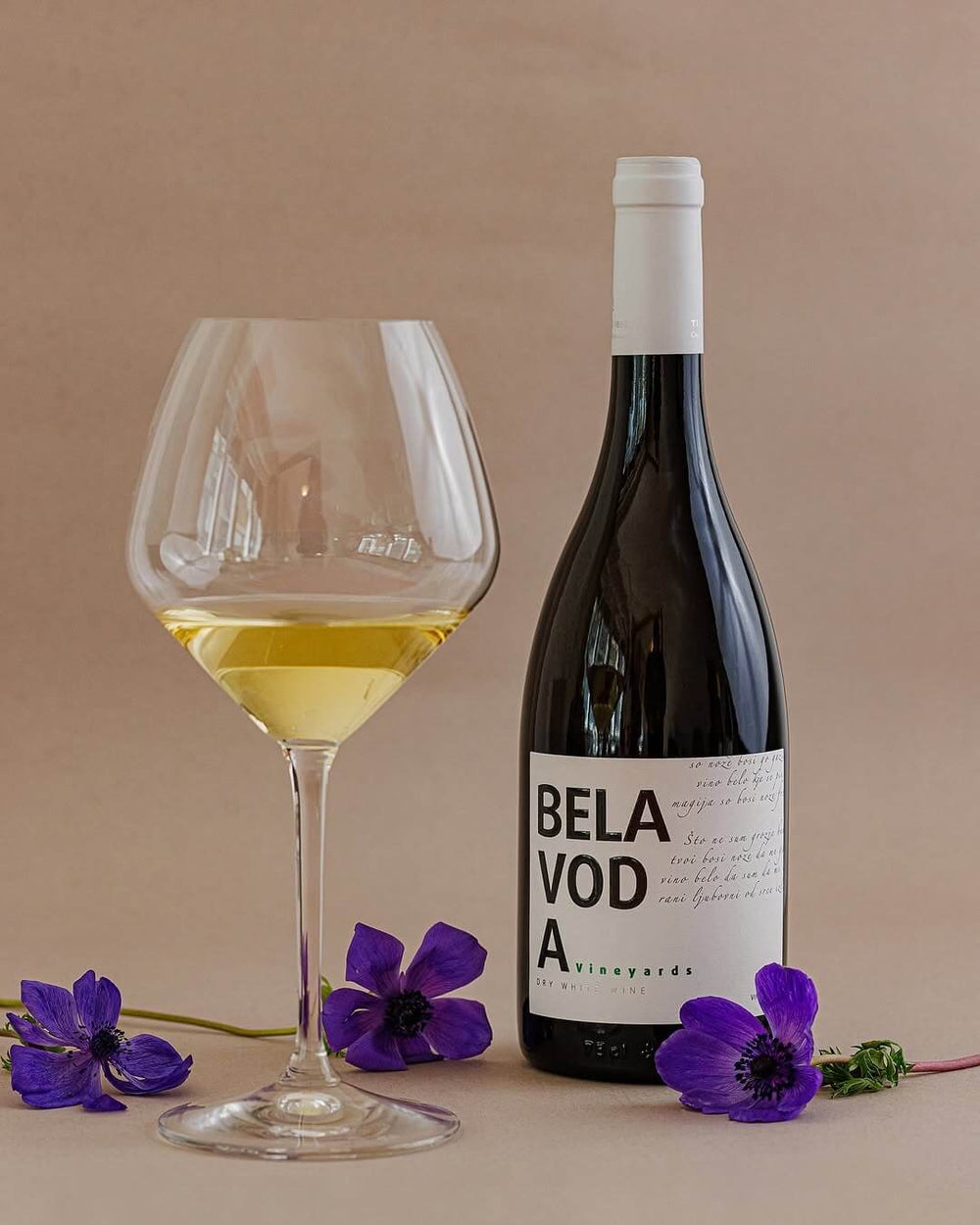 A bottle of Bela Voda White beside a glass of white wine and purple anemones