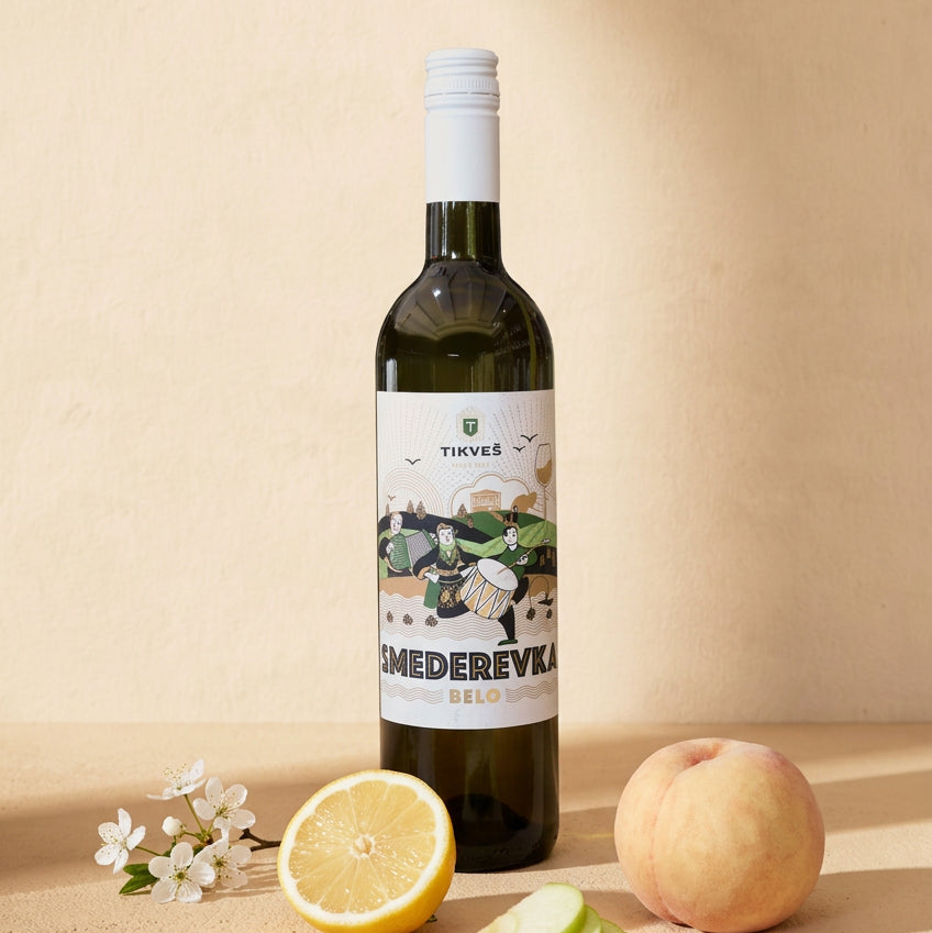 A bottle of Smederevka on a warm cream surface with half a lemon, green apple slices, a white peach and white blossom