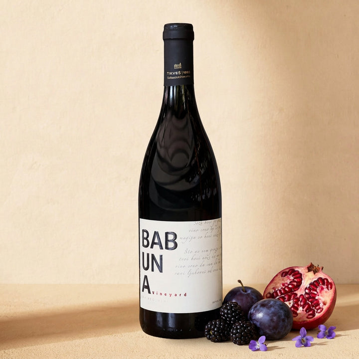 A bottle of Babuna Red on a warm cream surface with blackberries, dark plums, half a pomegranate and small violets