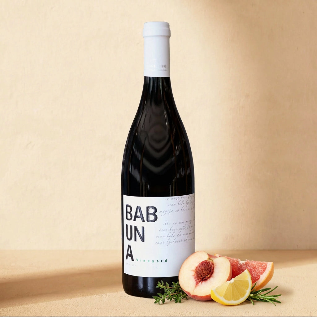 A bottle of Babuna White on a cream surface with a halved white peach, lemon, pink grapefruit and fresh herbs