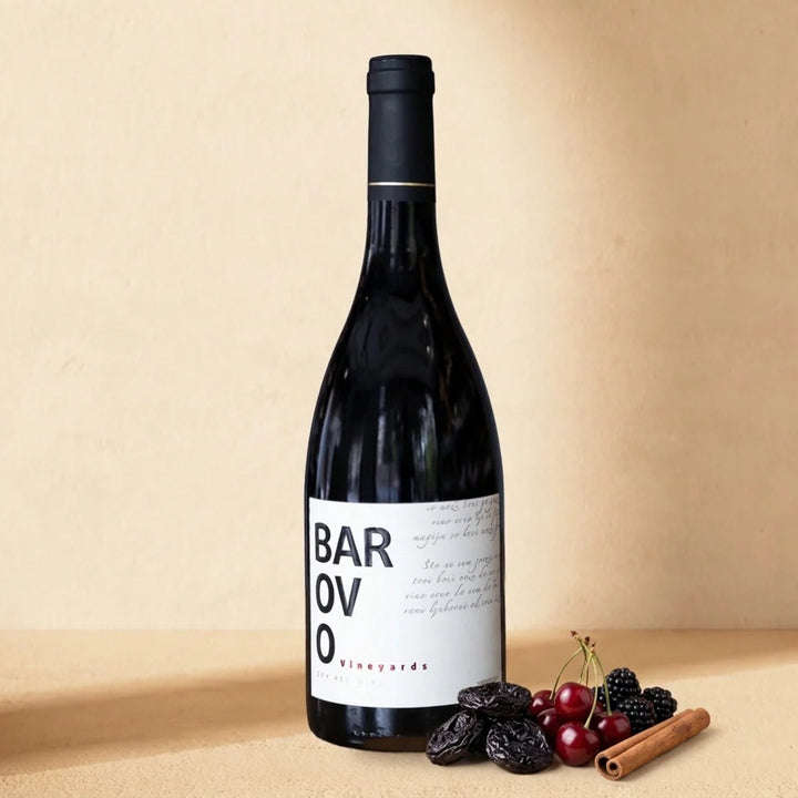 A bottle of Barovo Red on a warm cream surface with prunes, sour cherries, blackberries and a cinnamon stick