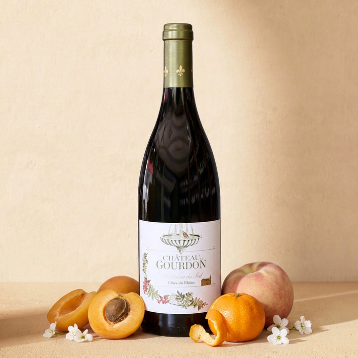 A bottle of Château de Gourdon Fontaine du Sud on a warm cream surface with halved apricots, a mandarin, a white peach and small white flowers