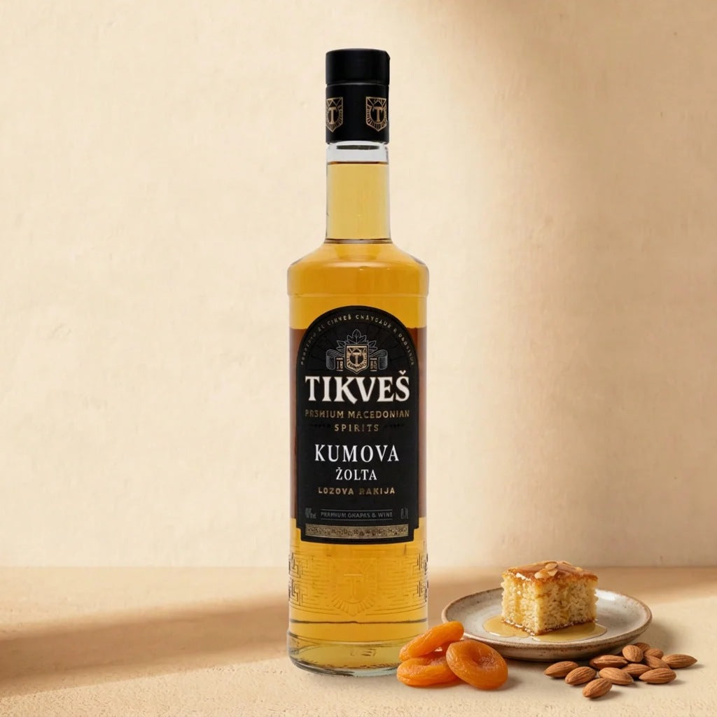 A bottle of Kumova Žolta rakija on a warm cream surface with a slice of almond honey cake, dried apricots and almonds