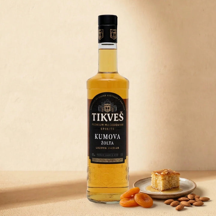 A bottle of Kumova Žolta rakija on a warm cream surface with a slice of almond honey cake, dried apricots and almonds
