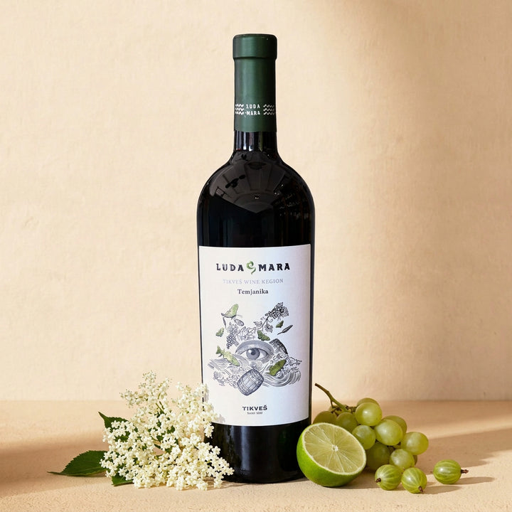 A bottle of Temjanika Luda Mara on a warm cream surface with elderflower, green grapes, half a lime and gooseberries