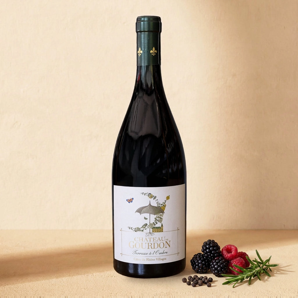 A bottle of Château de Gourdon Terrasse à l'Ombre on a warm cream surface with blackberries, raspberries, black peppercorns and rosemary