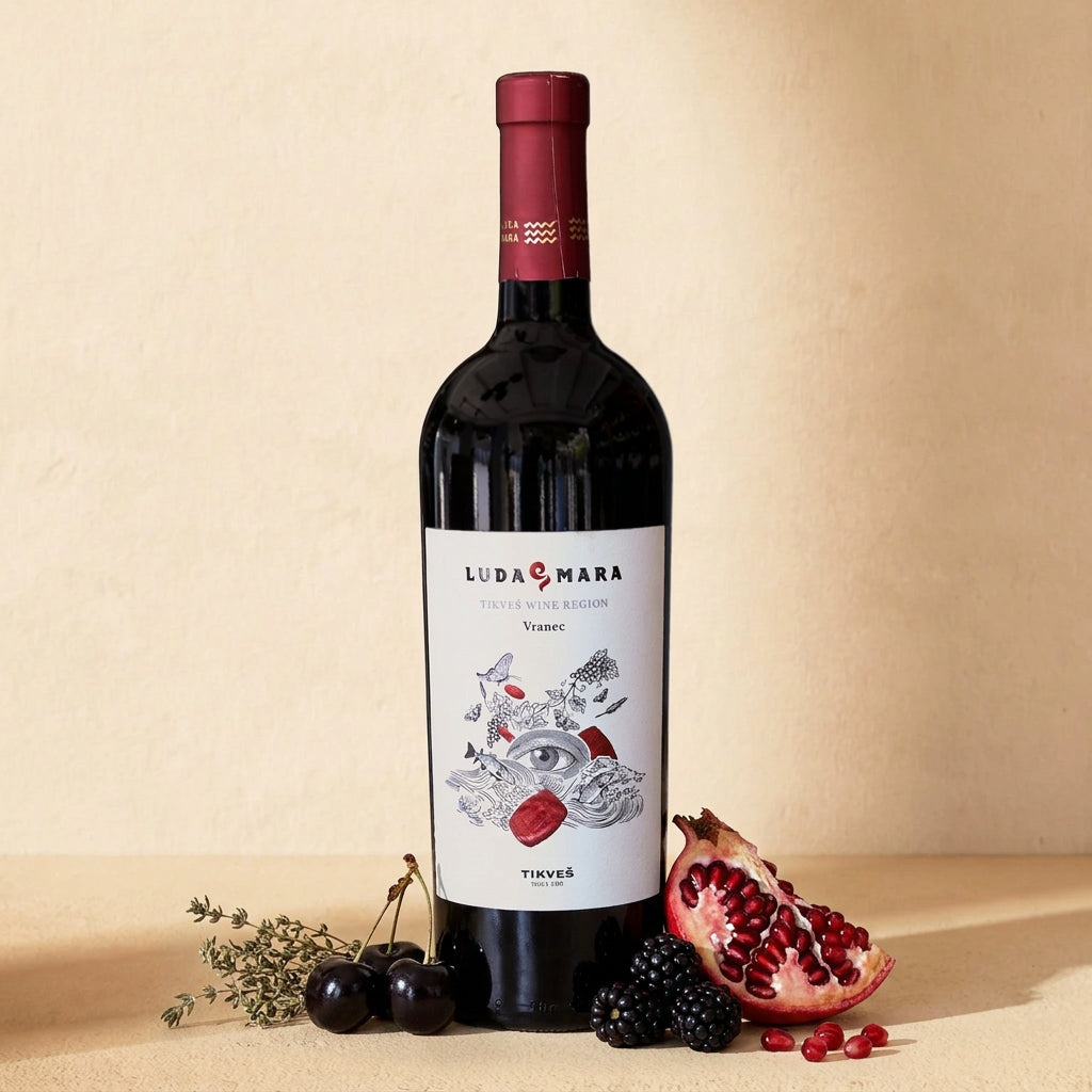 A bottle of Vranec Luda Mara on a warm cream surface with blackberries, black cherries, half a pomegranate and dried thyme
