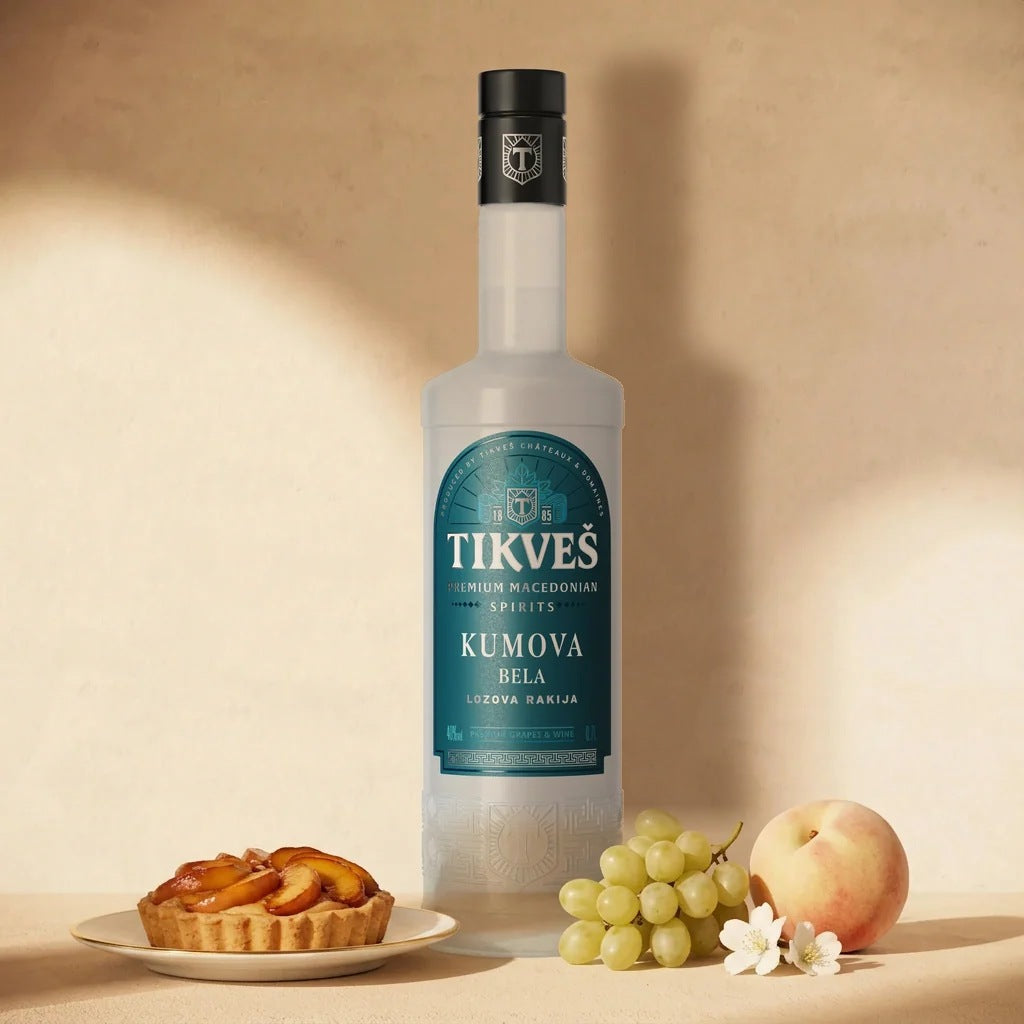A bottle of Kumova Bela rakija against a cream wall with a peach tart, muscat grapes, a white peach and small white flowers