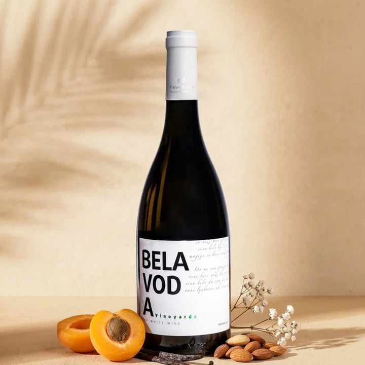 A bottle of Bela Voda White on a cream surface with halved apricots, a vanilla pod, almonds and dried white flowers